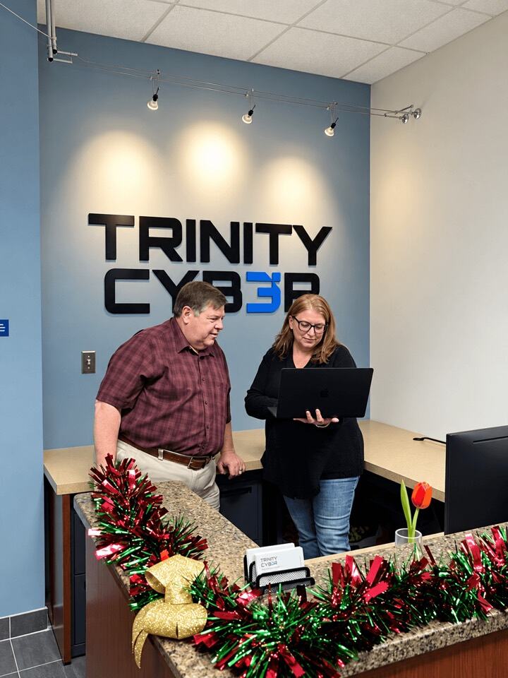 Two team members reviewing a laptop at the front desk beneath the Trinity Cyber sign
